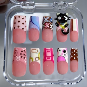 May include: A set of ten press-on nails in a clear plastic case, featuring a French manicure style with a nude base. Each nail has a unique design, including brown polka dots, blue swirls, floral patterns, a 3D black cat, and pink geometric shapes.