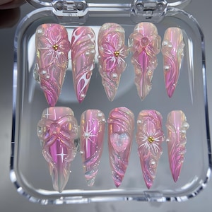 May include: A set of ten long, pointed artificial nails. Each nail is a translucent pink with intricate floral and pearl embellishments. Some nails feature a flower design with a gold center, while others have swirling patterns.