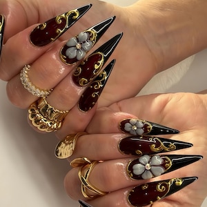 May include: Long stiletto-shaped artificial nails featuring a deep burgundy and black gradient base, accented with intricate gold filigree scrollwork and three-dimensional grey floral embellishments with small clear center crystals.