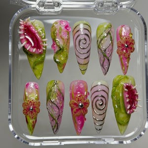 May include: A set of ten stiletto-shaped press-on nails in a clear plastic case. The designs feature pink and green botanical motifs, 3D sculpted Venus flytraps, raised floral accents with silver centers, and metallic silver spiral line patterns.