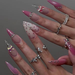 May include: A collection of long, stiletto-shaped acrylic nails in various designs. Some nails are solid pink, others feature a French tip, polka dots, or clear tips with bow accents. Several fingers display silver rings with sparkling embellishments.