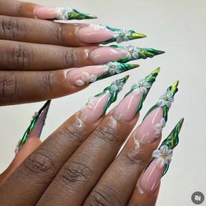 May include: Long, pointed fingernails with a pink base and white floral accents. The nails feature a green and gold design along the edges, creating a unique and artistic look. The nails are a form of nail art.