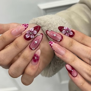 May include: A set of long, oval-shaped artificial nails. The nails feature a variety of designs, including a deep red base with white floral accents, a pink and red gradient, and a pink base with a red and white spotted design. Some nails have small, silver embellishments.