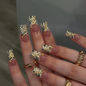 May include: Long, square-shaped acrylic nails with a zebra print design in black and cream, accented with gold studs. The nails feature a nude base and are adorned with gold rings.