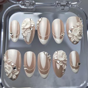 May include: A set of ten oval-shaped press-on nails. Each nail features a nude base with white and pearl accents. Some nails have 3D white rose embellishments and rhinestones. The nails are displayed in a clear plastic case.