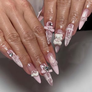 May include: Long, pointed fingernails with a light pink base and glitter accents. The nails are decorated with small bows, pearls, and a white teddy bear. Some nails have a clear, textured design.