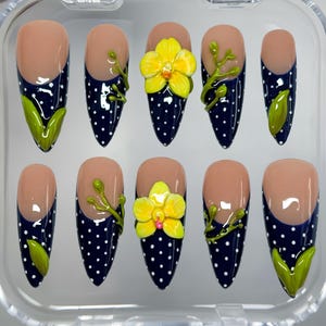 May include: Set of ten artificial nails with a nude base and navy blue tips with white polka dots. Some nails feature a 3D yellow orchid and green leaf accents. The nails are in a clear plastic case.