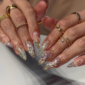 May include: Almond-shaped nails featuring intricate gold filigree patterns, iridescent base colors, and painted floral motifs in shades of pink and purple. The fingers are adorned with multiple gold rings, including bands with small embedded clear stones.