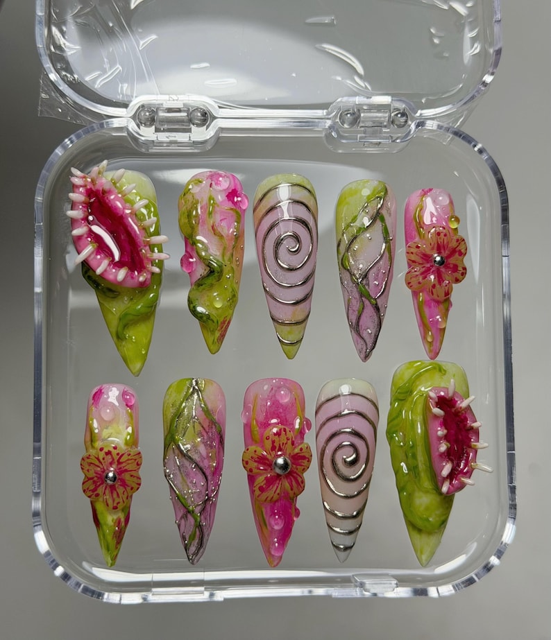 May include: A set of ten stiletto-shaped press-on nails in a clear plastic case. The designs feature pink and green botanical motifs, 3D sculpted Venus flytraps, raised floral accents with silver centers, and metallic silver spiral line patterns.