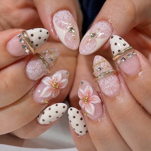 May include: Close-up of a set of manicured nails. The nails feature a variety of designs, including white lace patterns, pink bases, gold accents, and 3D flower embellishments. Some nails have a polka dot pattern.