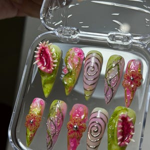 May include: A set of ten stiletto-shaped press-on nails in a clear plastic case. The designs feature 3D elements including pink Venus flytraps, raised silver swirls, textured green vines, and pink flowers with silver centers on a glossy base.