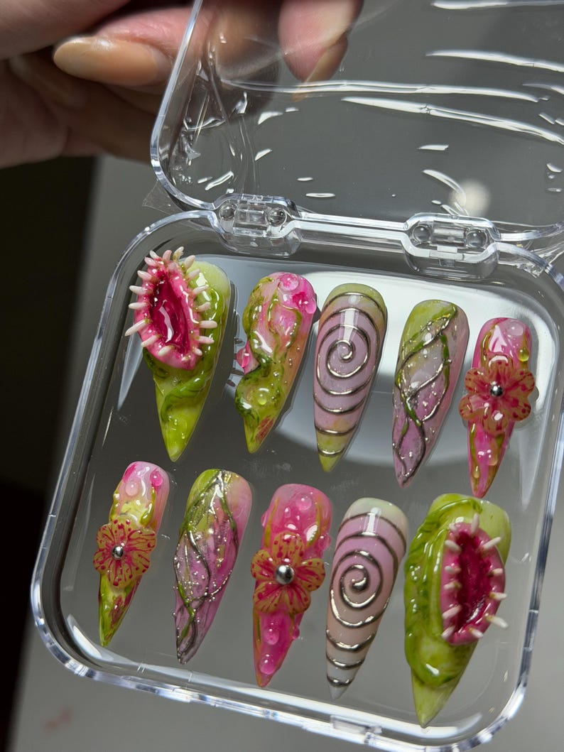 May include: A set of ten stiletto-shaped press-on nails in a clear plastic case. The designs feature 3D elements including pink Venus flytraps, raised silver swirls, textured green vines, and pink flowers with silver centers on a glossy base.