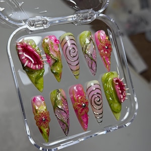May include: A set of ten stiletto-shaped press-on nails in a clear plastic case. The designs feature 3D sculpted Venus flytraps, pink and green floral patterns, silver spiral accents, and textured vines with small clear droplets on a glossy finish.