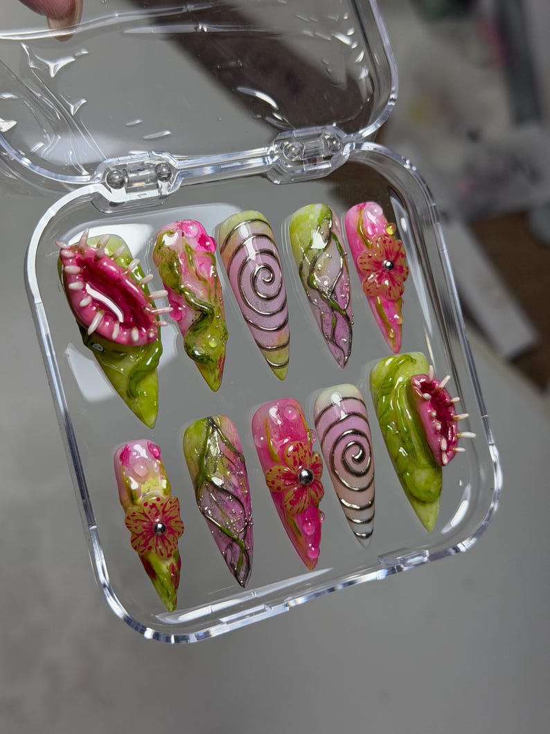 May include: A set of ten stiletto-shaped press-on nails in a clear plastic case. The designs feature 3D sculpted Venus flytraps, pink and green floral patterns, silver spiral accents, and textured vines with small clear droplets on a glossy finish.