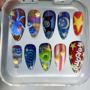May include: A set of ten artificial nails with various designs. The nails feature a variety of colors, including blue, red, yellow, and white. Some nails have 3D embellishments, such as stars and buttons. One nail has the word "Welcome" in white.