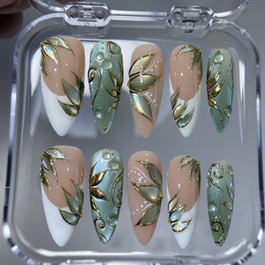 May include: A set of ten artificial nails in a clear plastic case. The nails feature a variety of designs, including white tips, nude bases, and light green and gold leaf accents. Some nails have water droplet details.