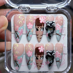 May include: A set of ten almond-shaped press-on nails in a clear plastic case. The design features a light blue and nude base with painted pink cherry blossoms, swirling yellow accents, brown branches, a cartoon character face, and a 3D black cat.