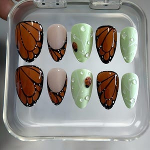May include: A set of ten artificial nails in a clear plastic case. The nails feature various designs, including monarch butterfly wing patterns in brown and black, light green leaves with ladybugs, and clear water droplet accents.