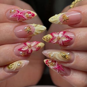 May include: Close-up of a set of manicured nails with a beach theme. Each nail features a light pink base with 3D designs of starfish, flowers, and seashells in yellow, pink, and gold. Small pearls and gold accents complete the look.