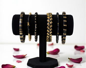 May include: A collection of black and gold bracelets displayed on a black velvet stand. The bracelets feature various designs, including beaded and patterned styles. Rose petals are scattered around the base, adding a touch of color.