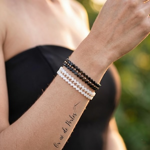 Könnte beinhalten: Drei Perlenarmbänder in Schwarz, Weiß und Gold werden an einem Arm gezeigt. Die Armbänder haben ein Zickzackmuster. Der Arm hat ein Tattoo mit der Aufschrift "Or né de Perles". Die Person trägt ein schwarzes trägerloses Oberteil.
