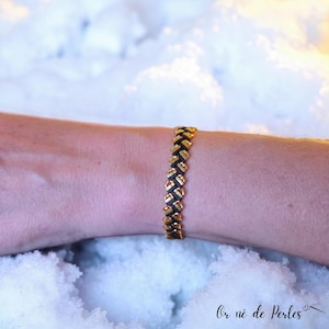Peut inclure: Un bracelet doré et noir avec un motif tissé. Le bracelet est composé de petits maillons rectangulaires dorés et de maillons noirs. Le bracelet est porté au poignet d'une personne. L'arrière-plan est une scène enneigée floue.
