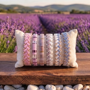 Op de afbeelding: Zes kralenarmbanden gerangschikt op een klein rechthoekig beige stoffen kussen. De armbanden hebben verschillende patronen, waaronder visgraatvlechten en gestapelde rechthoekige kralen in tinten roze, mauve, lavendel, wit en grijs met gouden accenten.