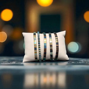 May include: A collection of beaded bracelets in blue, gold, and brown hues are presented on a white cushion. The bracelets feature diverse designs, including rectangular and round beads. The background is blurred with bokeh lights.