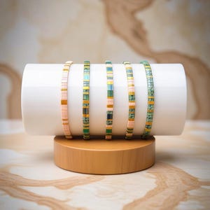 Peut inclure: Cinq bracelets de perles de différentes couleurs, présentés sur un support cylindrique blanc sur une base en bois. Les bracelets présentent un mélange de perles rectangulaires roses, vertes, bleues et jaunes. Le fond a un motif de type marbre.