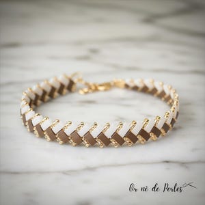 Peut inclure: Un bracelet à motifs en chevrons avec des perles rectangulaires blanches et marron alternées, reliées par des accents dorés. Le fermoir et la chaîne sont également dorés. Le bracelet est présenté sur une surface en marbre. Le texte "Or né de Perles" est visible.