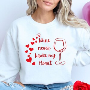May include: White sweatshirt with the text "Wine never broke my Heart" in red, along with a wine glass and red heart accents. The sweatshirt is part of a Valentine's Day theme.