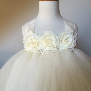 May include: A white tulle dress with a lace bodice and three large white fabric flowers. The dress is on a white mannequin.