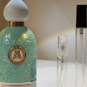 Pu&ograve; includere: Un flacone di profumo turchese con tappo bianco e oro, un'emblema dorato con la lettera "A" e le parole "SLATE BLUE". Due flaconi spray in vetro trasparente sono accanto al flacone di profumo.