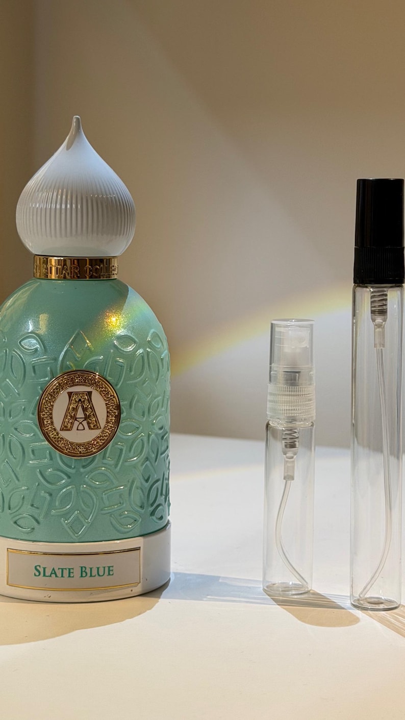 Pu&ograve; includere: Un flacone di profumo turchese con tappo bianco e oro, un'emblema dorato con la lettera "A" e le parole "SLATE BLUE". Due flaconi spray in vetro trasparente sono accanto al flacone di profumo.