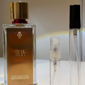 May include: A rectangular perfume bottle with a gold cap and a gold-colored label that reads "TILIA" and "MARC-ANTOINE BARROIS PARIS." Two clear, empty spray bottles are next to it.