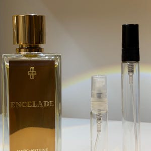 Pode incluir: Um frasco de perfume retangular com tampa dourada e rótulo dourado que diz "ENCELADE" e "MARC-ANTOINE BARROIS PARIS". Dois frascos de vidro transparente com borrifadores estão ao lado do frasco. O frasco de perfume tem um emblema dourado.