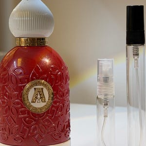Pode incluir: Um frasco de perfume vermelho com uma tampa branca e dourada, um emblema dourado com a letra "A" e a palavra "QALBI" no rótulo. Dois frascos de spray de vidro transparente estão ao lado do frasco de perfume.