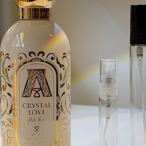 Peut inclure: Un flacon de parfum blanc avec des détails dorés et un bouchon doré, intitulé "CRYSTAL LOVE for her". Deux flacons vaporisateurs transparents et vides sont à côté. Le flacon de parfum est de 100 ml.