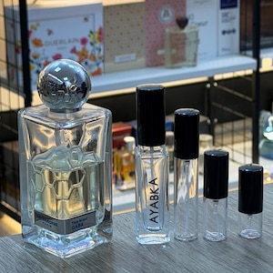 Pode incluir: Uma coleção de frascos de perfume e atomizadores. Um frasco de perfume grande e quadrado com uma tampa prateada e um design de favo de mel está ao lado de atomizadores menores e transparentes com tampas pretas. Um atomizador está rotulado como "AYABKA".