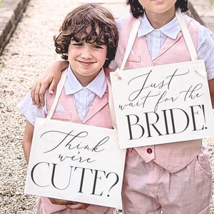 May include: Two young boys in pink suits holding signs that say "Think we're cute?" and "Just wait for the bride!" They are standing on a stone path in front of a house.