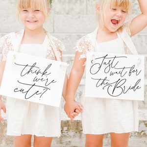 May include: Two young girls wearing white dresses and flower crowns hold signs with calligraphy text. One sign reads "think we're cute?" and the other says "Just wait for the Bride". They are standing on stone steps, holding hands, and smiling.