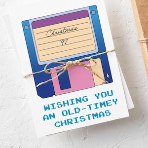 May include: A bundle of Christmas cards tied with string, each with a retro floppy disk design. The cards read "Wishing you an old-timey Christmas" and "Christmas '97". A wrapped gift box with pinecones and greenery is next to the cards.