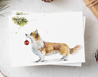 May include: A white greeting card featuring a watercolor illustration of a brown and white Corgi dog holding a small red spherical Christmas ornament in its mouth, with a green pine sprig painted above the dog's head.