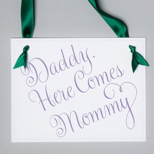 May include: White rectangular sign with the text "Daddy, Here Comes Mommy" in purple script. The sign is suspended by a green satin ribbon. Small green ribbon accents are tied on the corners.