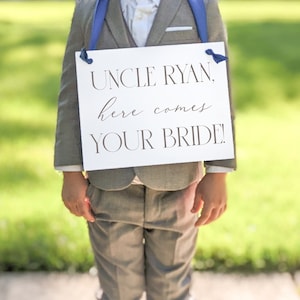 May include: A young boy in a gray suit holds a sign that says "Uncle Ryan, here comes your bride!" The sign is white with black lettering and has blue ribbon ties.