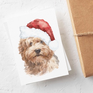 May include: Watercolor Christmas cards featuring a brown and white dog wearing a Santa hat. A wrapped brown gift box with twine, pine sprigs, and pine cones is next to the cards. The cards and gift box are on a white surface.