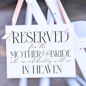 May include: White chair with a sign that reads "RESERVED for the MOTHER of the BRIDE who is celebrating with us IN HEAVEN". The sign is attached to the chair with a pink ribbon.