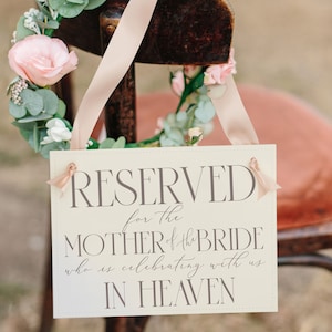 May include: A white sign with the text "RESERVED for the MOTHER of the BRIDE who is celebrating with us IN HEAVEN" hanging on a wooden chair decorated with a floral wreath.
