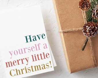 May include: A white greeting card with the text Have yourself a merry little Christmas! in teal, pink, red, and gold lettering, placed next to a brown paper-wrapped gift decorated with pine needles and three small brown pinecones.