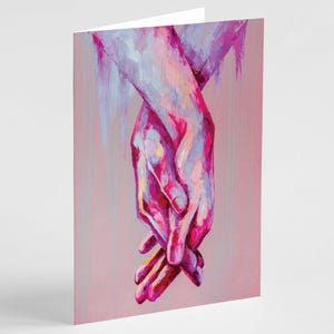 May include: Greeting card featuring an artistic painting of two hands clasped together. The artwork uses a palette of pink, purple, and blue hues, with a soft, painterly style. The card is on a pink background.